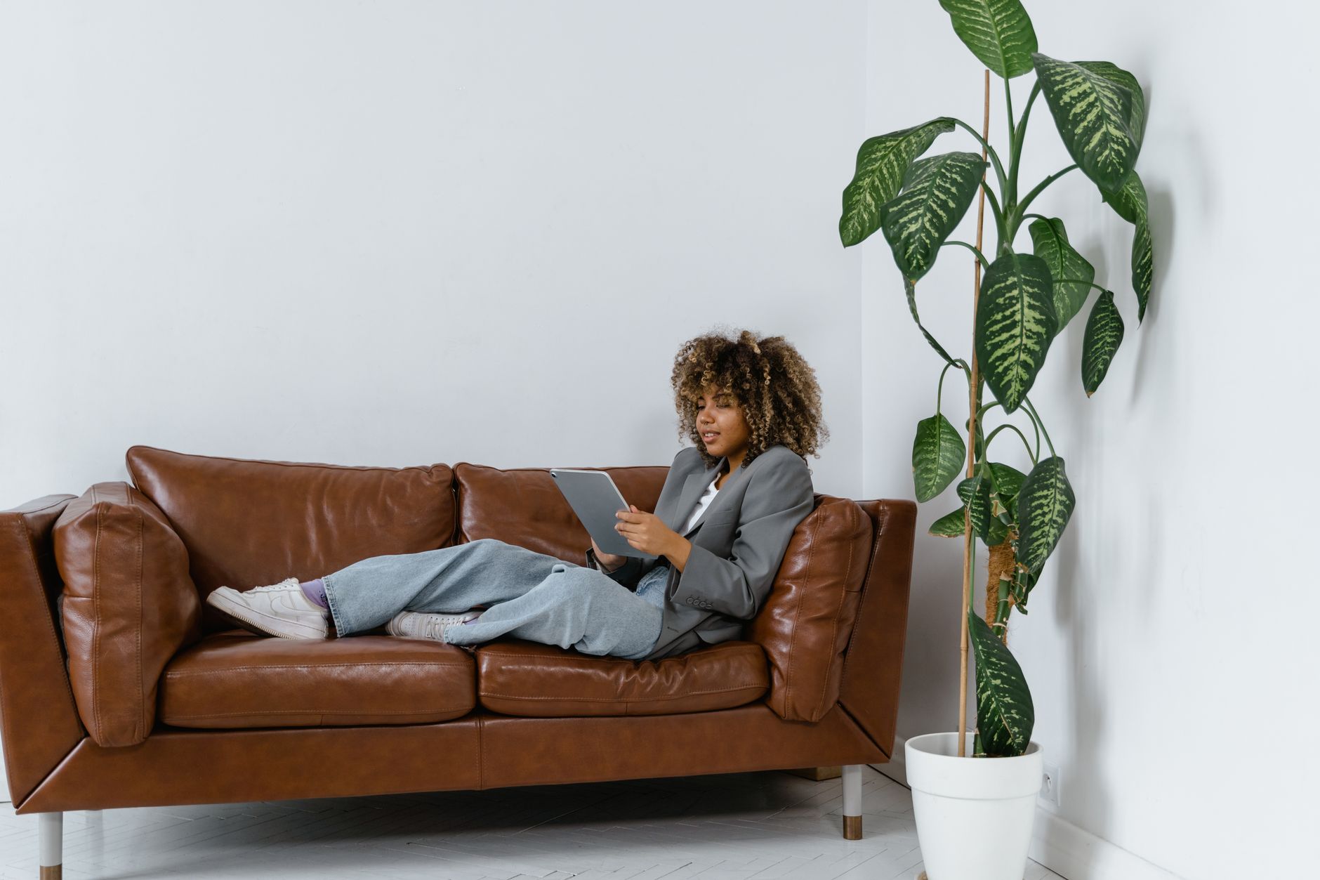 Mulher afro-americana relaxada num sofá de pele em casa, com um tablet e uma planta ao lado.