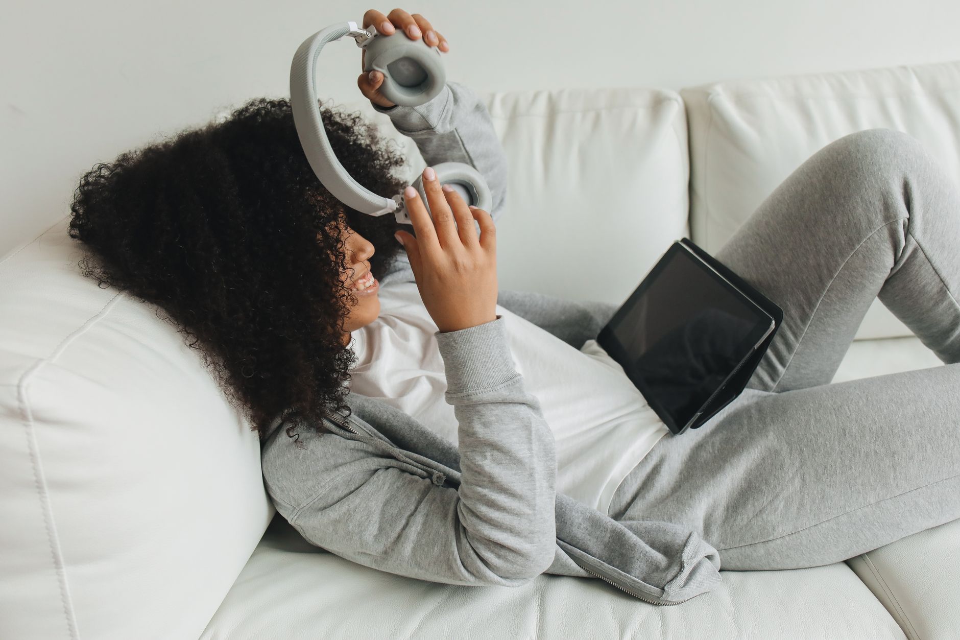 Mulher negra com auscultadores, relaxada num sofá em casa com um tablet.